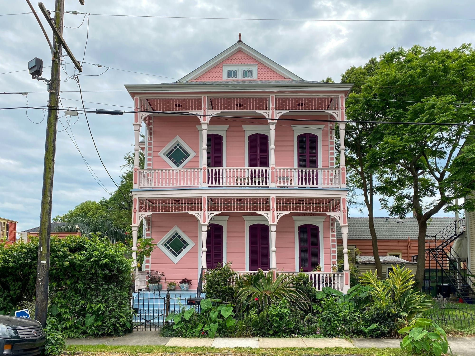 A tall pink house with two porches, white trim, and purple shutters, surrounded by lush green plants.