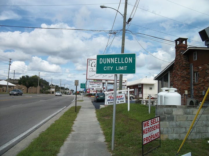 A roadside “Dunnellon City Limit” sign stands beside a busy street with shops and cars.