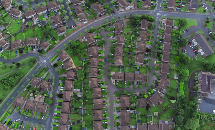 An aerial view shows a neatly arranged suburban neighborhood with rows of houses, tree-lined streets, green lawns, and parked cars forming a quiet residential grid.