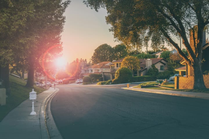 A calm, tree-lined suburban street curves past tidy homes as the low sun flares warmly through the branches at sunset.