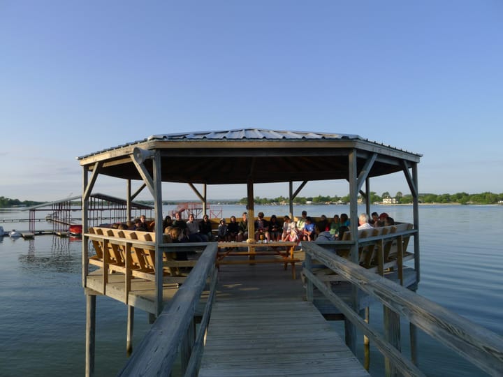 A diverse group sits on wooden benches under an open pavilion on a lake dock, gathered for an outdoor event at sunset, with calm water, distant shoreline, and boats in the background.
