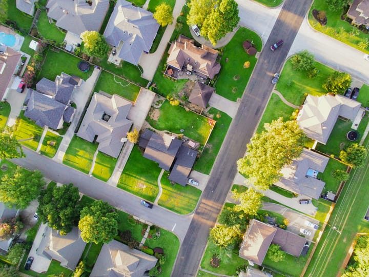 An aerial view of a quiet suburban neighborhood with neatly spaced houses, green lawns, trees, and intersecting streets.