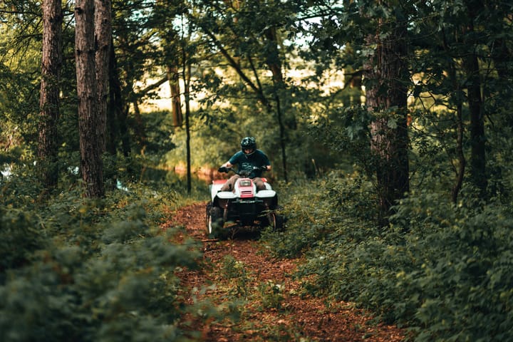 A person wearing a helmet rides a four-wheeler along a narrow dirt trail through a dense, green forest.