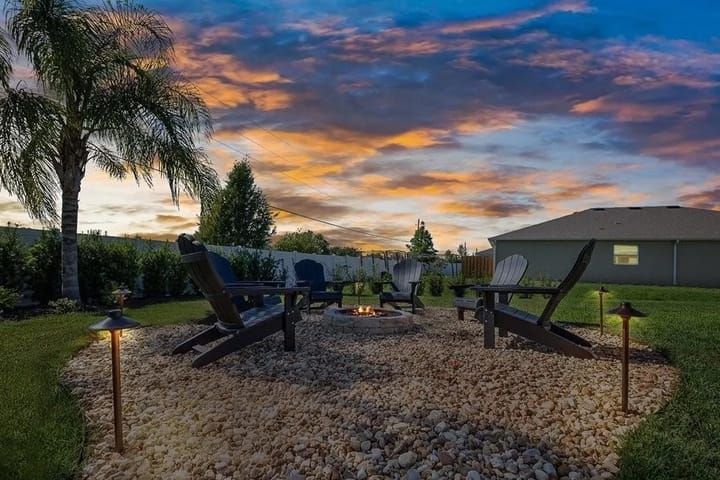 A backyard fire pit surrounded by chairs sits under a colorful sunset sky with palm trees nearby.