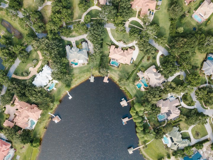 An aerial view shows large homes with pools arranged around a small lake, surrounded by green lawns and trees.