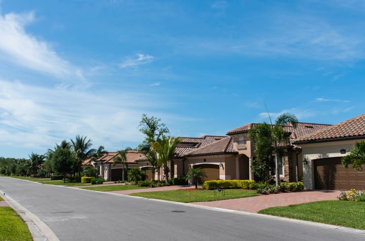 A sunny Florida suburban street lined with upscale Mediterranean-style homes, manicured lawns, and palm trees.