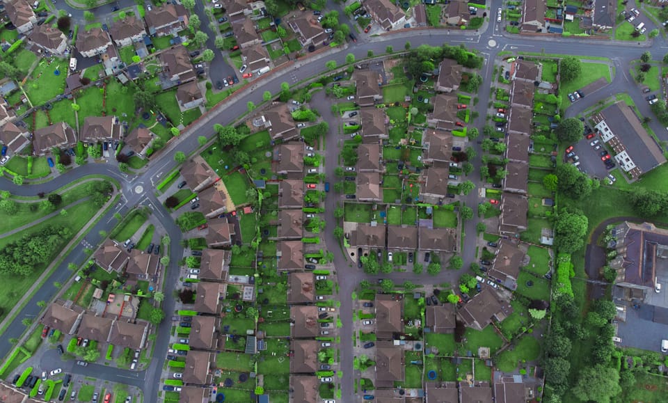 An aerial view shows a neatly arranged suburban neighborhood with rows of houses, tree-lined streets, green lawns, and parked cars forming a quiet residential grid.
