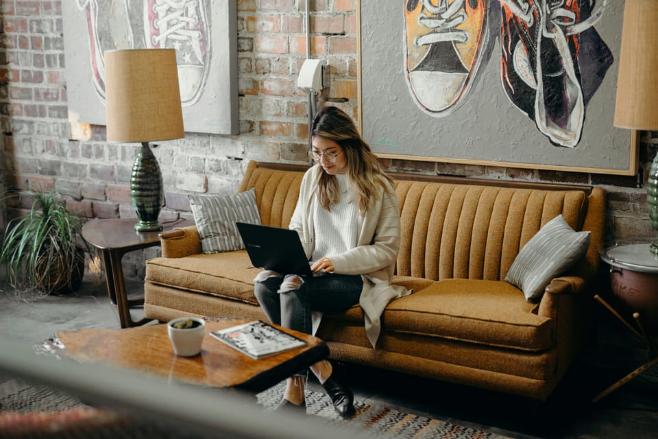 A young woman with glasses works on her laptop while sitting on a mustard-yellow sofa in a trendy, eclectic space.