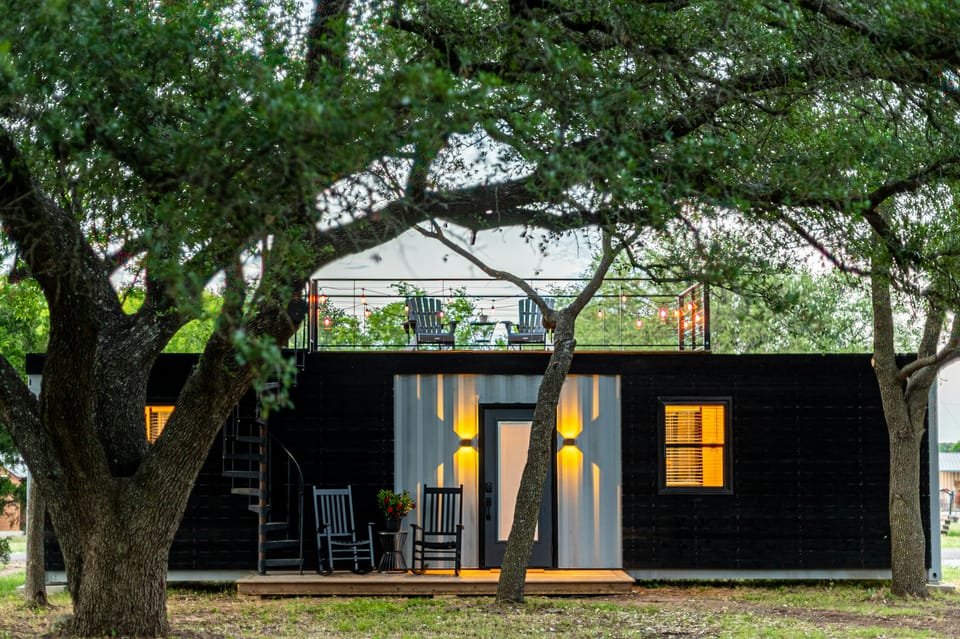 A glowing black container home with rocking chairs and a rooftop deck, framed by oak trees at dusk.