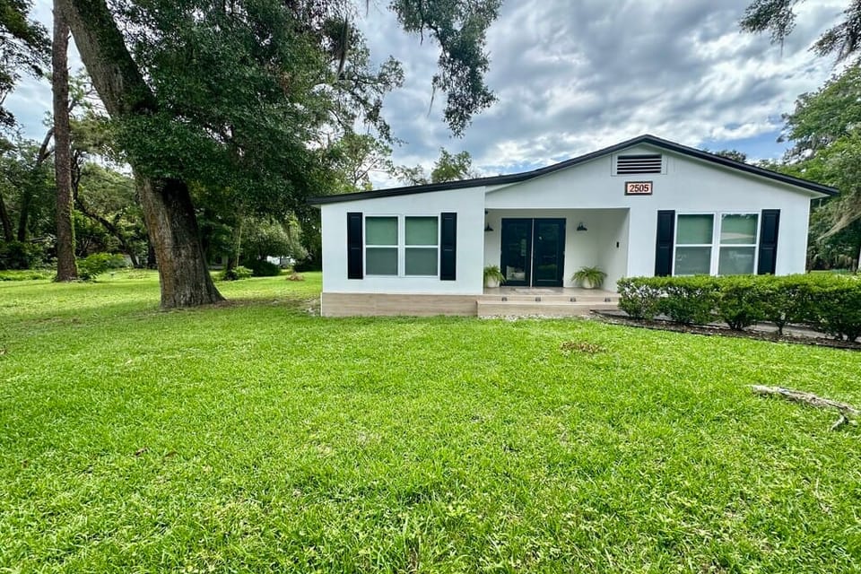 A white house with dark shutters sits on a green lawn under trees and cloudy skies.